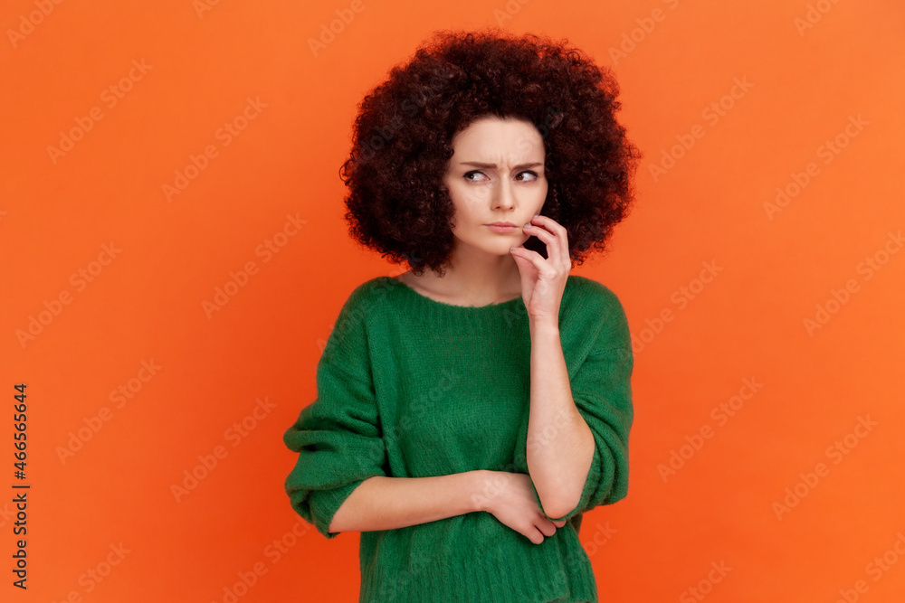 Pensive woman with Afro hairstyle wearing green casual style sweater thinking, looking away, having puzzled confused facial expression. Indoor studio shot isolated on orange background. Pensive woman with Afro hairstyle wearing green casual style sweater thinking, looking away, having puzzled confused facial expression. Indoor studio shot isolated on orange background.
