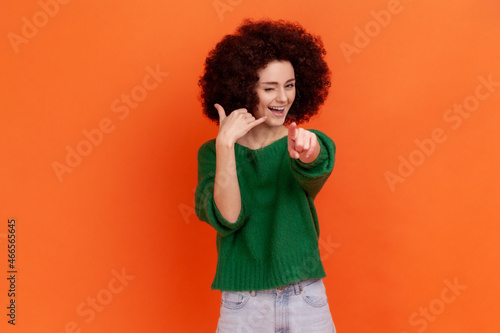 Hey you, call me! Woman with Afro hairstyle standing with telephone hand gesture and smiling to camera, flirting offering to contact by phone. Indoor studio shot isolated on orange background.