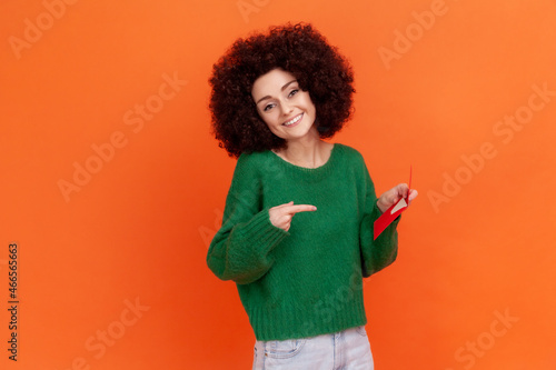 Smiling woman with Afro hairstyle wearing green casual style sweater holding and pointing at red envelope in her hand, romantic letter. Indoor studio shot isolated on orange background.