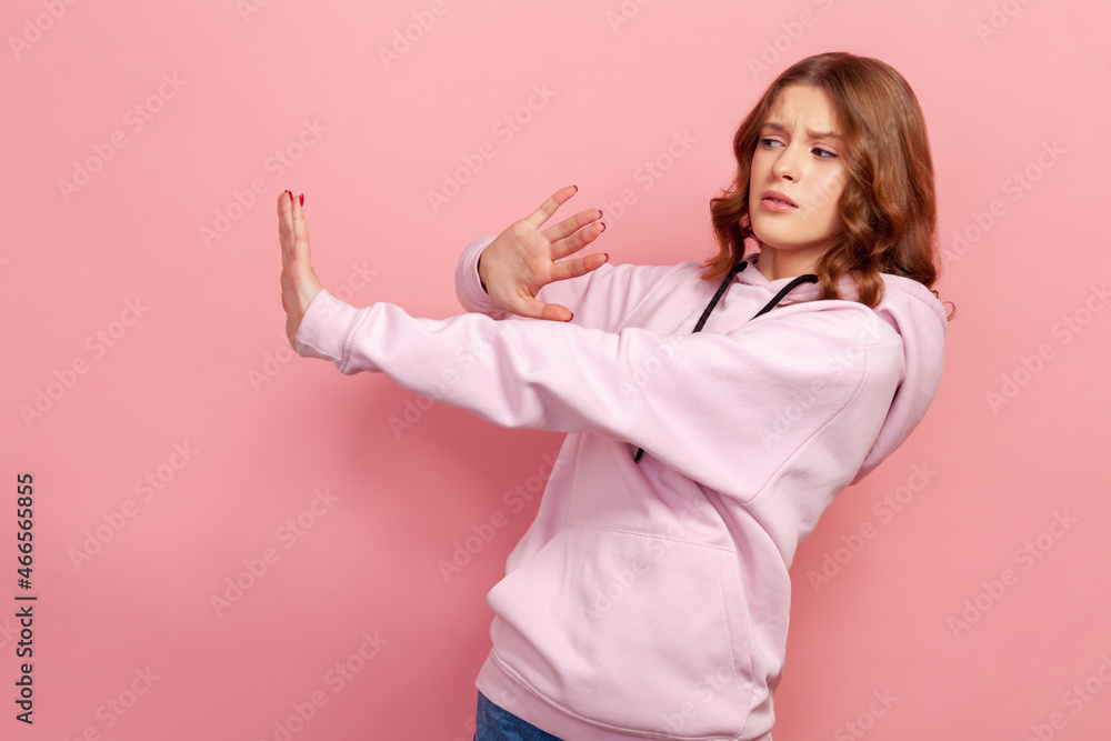 Side view of scared young girl standing with raised hands, gesturing stop, panicking looking frightened of danger, phobia. Indoor studio shot isolated on pink background Side view of scared young girl standing with raised hands, gesturing stop, panicking looking frightened of danger, phobia. Indoor studio shot isolated on pink background