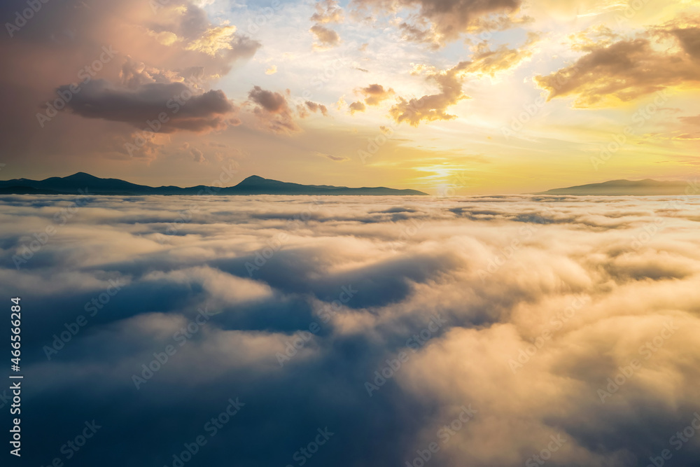 Aerial view of vibrant sunset over white dense clouds with distant dark mountains on horizon. Aerial view of vibrant sunset over white dense clouds with distant dark mountains on horizon.