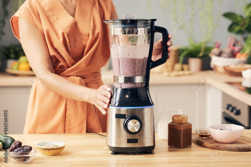 Hand of young woman preparing fruit smoothie in electric blender while standing by kitchen table photo