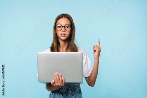 Smart teenager schoolgirl holding laptop computer showing pointing on copy space having idea, surfing webpages on Internet, social media posting,e-learning remotely isolated in blue background © InsideCreativeHouse