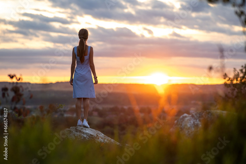 Dark silhouette of a young woman in summer dress standing outdoors enjoying view of nature at sunset. © bilanol