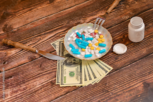 A plate of various medicinal pills is on dollar bills. The knife and fork are on the plate. The concept of expensive medical care.