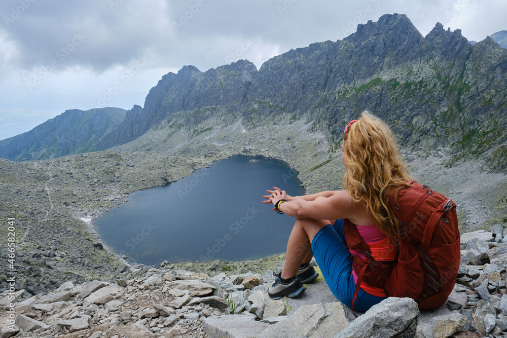 Woman hiker with curly blonde hair and red backpack looks towards Vysne Wahlenbergovo lake in Slovakia High Tatras mountains. Hiking, trekking, tourism, active, vacation, overcast.