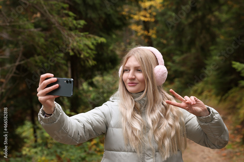 Young woman in warm earmuffs taking selfie near forest on autumn day
