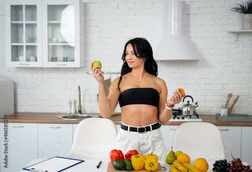 This or this. Pretty brunette caucasian woman standing near table with healthy food and making choice while looking at the green apple. Healthy diet food concept