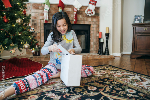 Slika na platnu Excited happy girl, unwrapping present in front of Christmas tree and fireplace