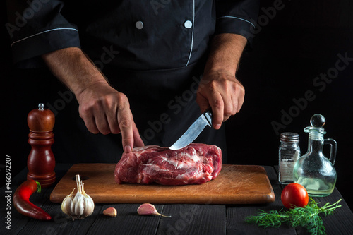 The cook cuts meat with a knife in the kitchen prepares food. Vegetables and spices on kitchen table in a restaurant to prepare a delicious lunch