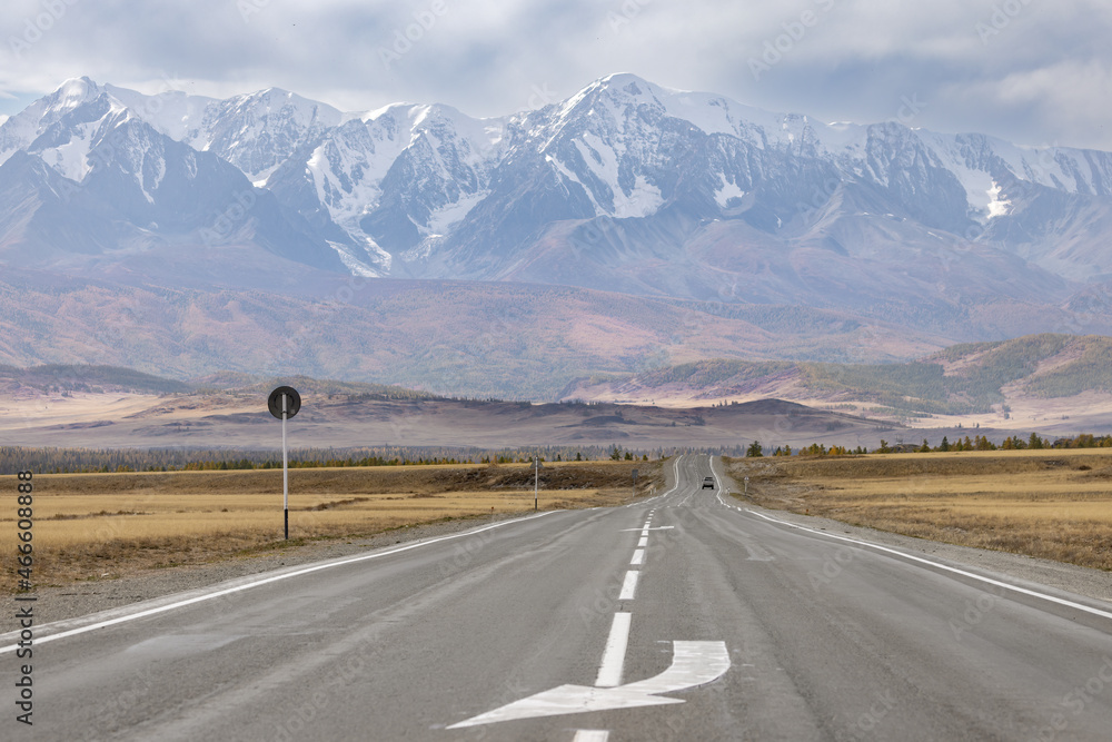 A road with markings in a mountain valley on the background of a mountain range in autumn. There's a car in the background. A road with markings in a mountain valley on the background of a mountain range in autumn. There's a car in the background.