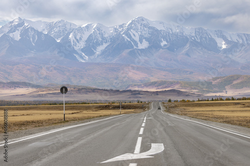 A road with markings in a mountain valley on the background of a mountain range in autumn. There's a car in the background.