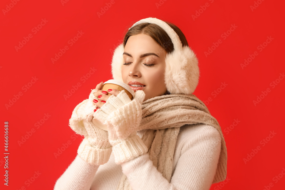 Beautiful young woman in warm earmuffs drinking coffee on red background Beautiful young woman in warm earmuffs drinking coffee on red background