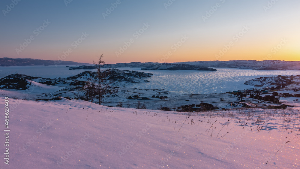 Dawn over the frozen Siberian lake. The sky is highlighted in orange. The glare of the sun on the ice. Sparkling snow in the foreground. The golden hour. Baikal