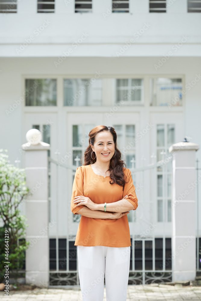 Happy smiling mature woman standing at gates of her mansion with arms crossed Happy smiling mature woman standing at gates of her mansion with arms crossed