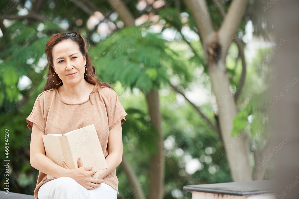 Naklejka premium Pensive mature woman sitting in park with opened book thinking after finishing reading