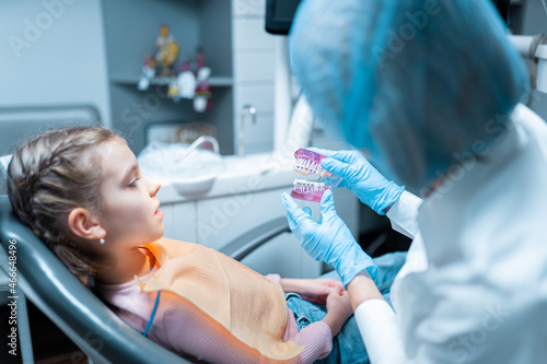 Friendly female dentist orthodontist educating little girl patient using teeth model with brecets.