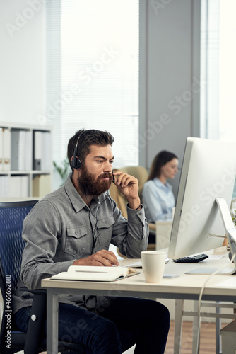 Serious young male helpdesk operator with beard adjusting headset microphone and answering call of client in office © DragonImages