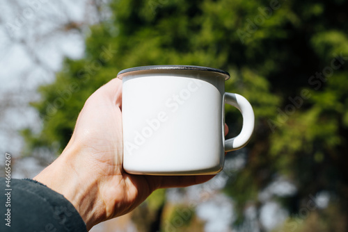 Concept hike, camping, trip. Hand showing empty white enamel mug with blank space for text or branding against forest outdoors background. Travel cup mock up