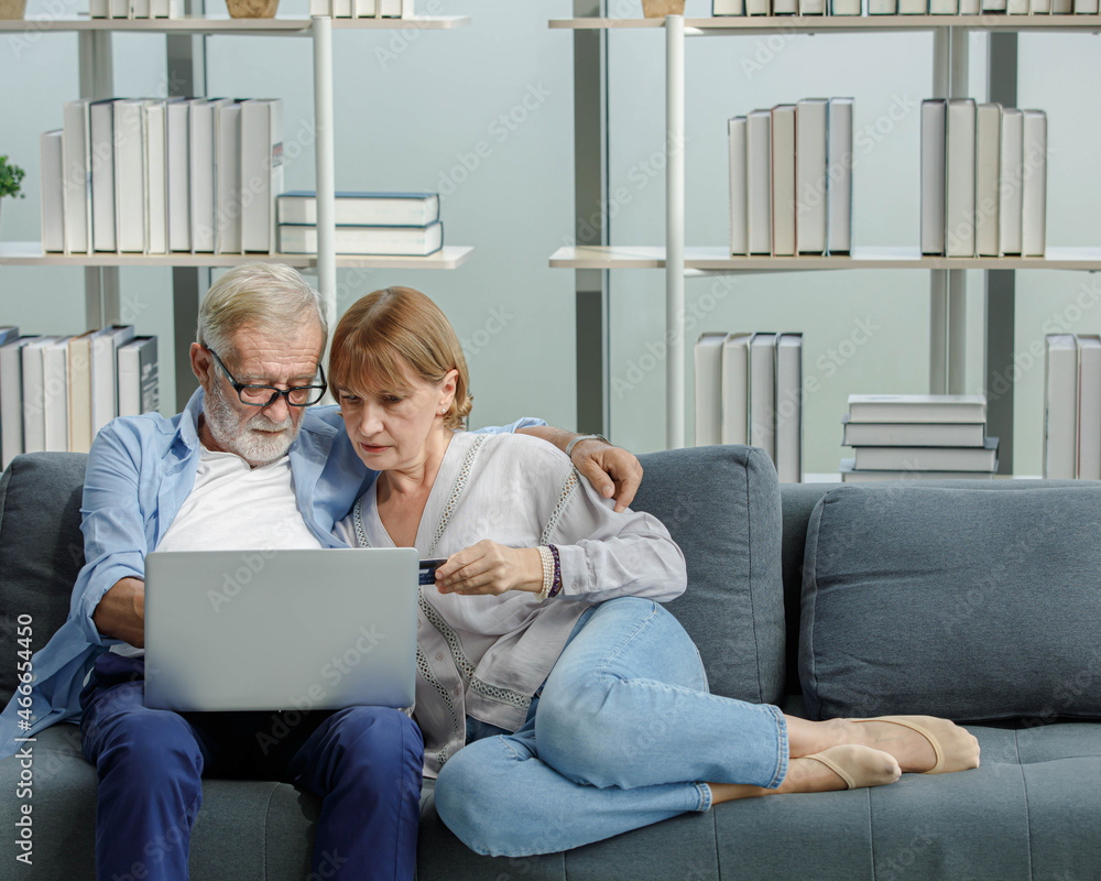 Caucasian old senior elderly grandparents couple sitting cuddling hugging together on sofa in living room at home using credit card and laptop computer browsing internet shopping from online store