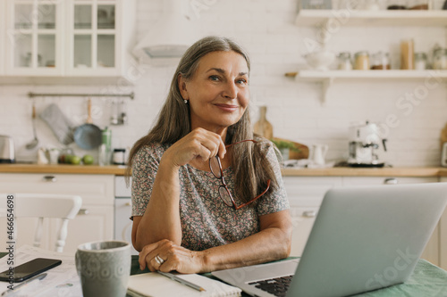 Picture of mature teacher holding glasses in hand, looking at camera with cute smile working on her laptop online from her kitchen, checking test results of her students, copybook, mug, pen on table