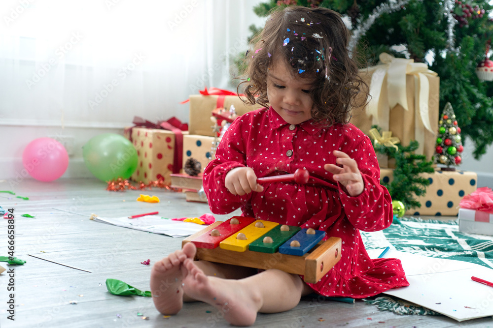 Cute little girl play wooden toy xylophone on floor, Christmas tree and gift boxes background,