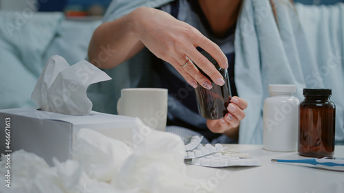 Close up of woman reading labels of tablets and jars with pills and capsules on table with medicaments, tissues and thermometer. Sick person looking for treatment to cure virus symptoms