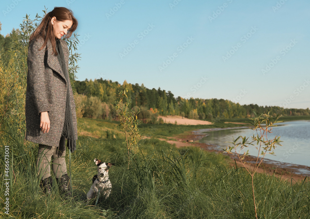 happy young woman walking with her dog along river wearing autumn outfit. caucasian woman with long hair looking at jack russell terrier pet and smiling. companion dog.