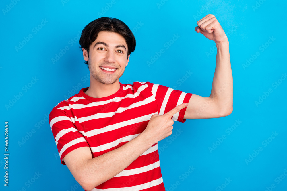 Photo of pretty confident young man wear striped t-shirt pointing finger arm muscle smiling isolated blue color background Photo of pretty confident young man wear striped t-shirt pointing finger arm muscle smiling isolated blue color background