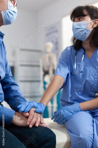 Nurse with face mask and gloves supporting senior man with orthopedic injury after osteopathy checkup in office. Medical assistant talking to old patient during coronavirus pandemic