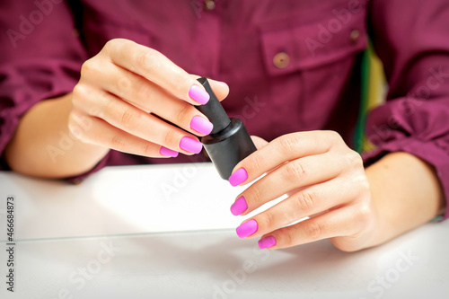 White woman holding nail polish black bottle with painted pink nails close up.