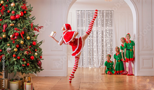 Children dressed as elves watch a dancing santa claus ballerina near a Christmas tree in a spacious white studio. photo