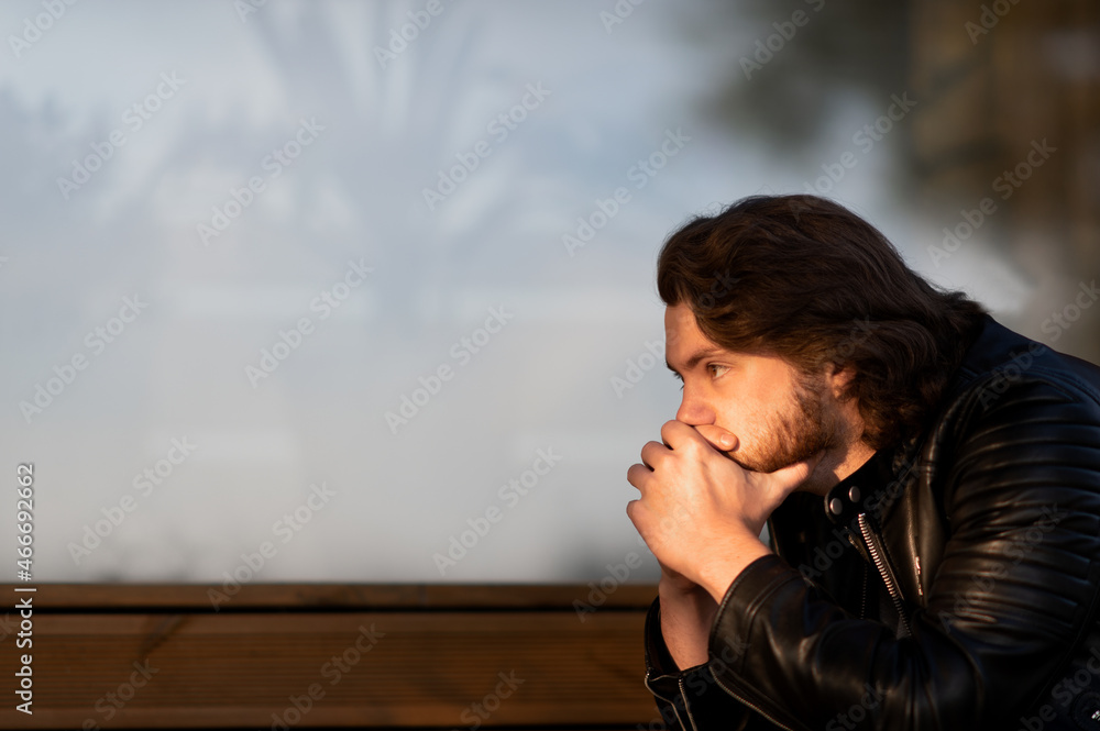 Fototapeta premium Pensive serious young bearded man wear black leather jacket sitting and thinking outdoors
