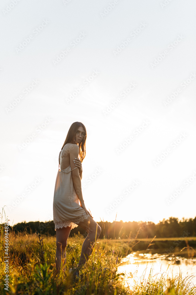 Obraz premium a young beautiful girl, of model appearance, in a shirt, stands among the landscape on the river bank, the setting sun illuminates her figure