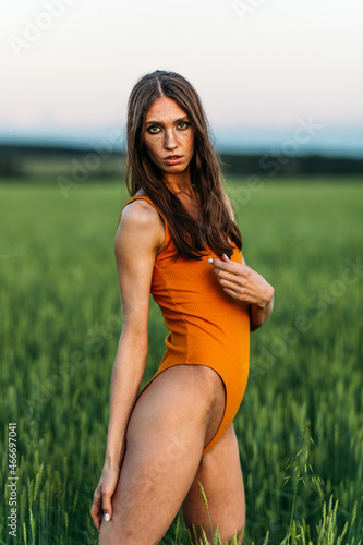 a young beautiful girl of model appearance in a bright orange bodysuit stands in the middle of a lush green field.