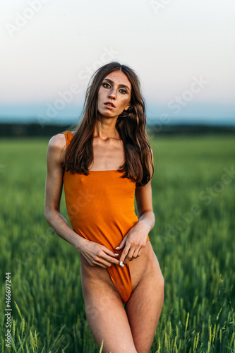 a young beautiful girl of model appearance in a bright orange bodysuit stands in the middle of a lush green field.
