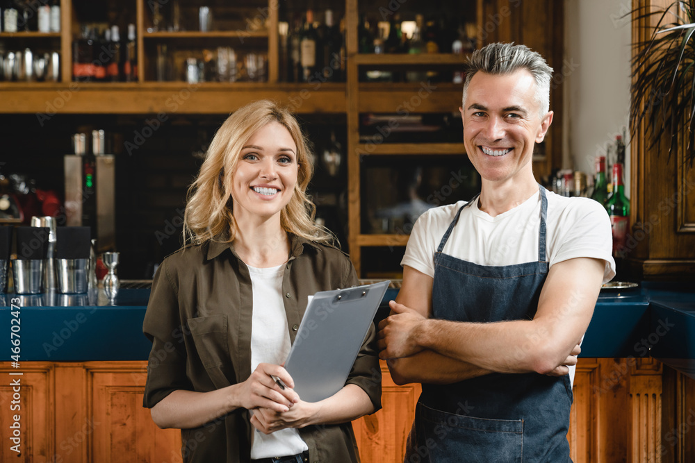 Smiling caucasian small business owner waiter barista with female auditor holding clipboard standing at the bar restaurant cafe counter checking products quality looking at the camera - obrazy, fototapety, plakaty 