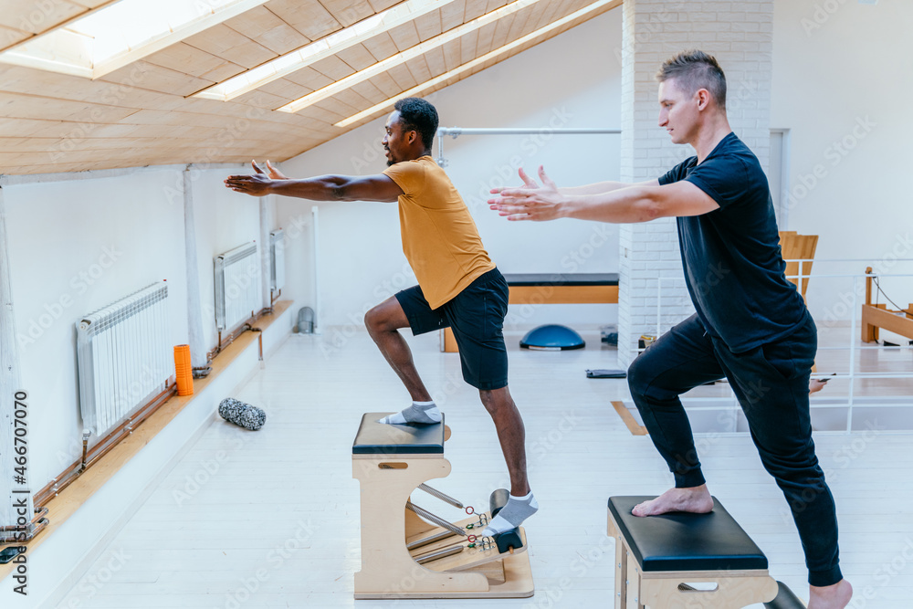 Fototapeta premium African american man doing exercise on pilates chair equipment. Caucasian male personal trainer helping him. Workout in rehabilitation center, pilates studio. Balancing herself with a bent leg.