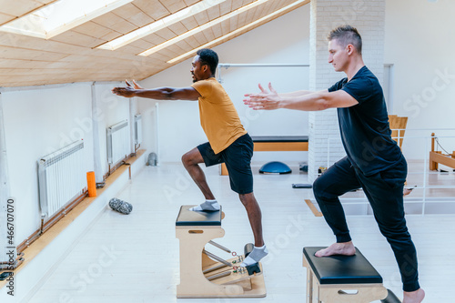African american man doing exercise on pilates chair equipment. Caucasian male personal trainer helping him. Workout in rehabilitation center, pilates studio. Balancing herself with a bent leg. © Iryna