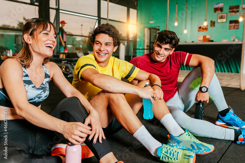 Portrait of a group of smiling friends laughing together after a gym class sitting on the floor. Drinking isotonic drinks and recovering after exercise. Healthy lifestyle concept.