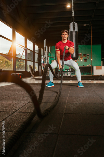 Athletic young man with battle rope doing exercise in functional training fitness gym, in day time with sunlight at window. Powerful fit athlete training.