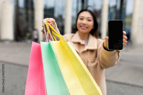 Happy young Asian woman showing shopper bags and smartphone with empty screen near big mall outdoors, mockup