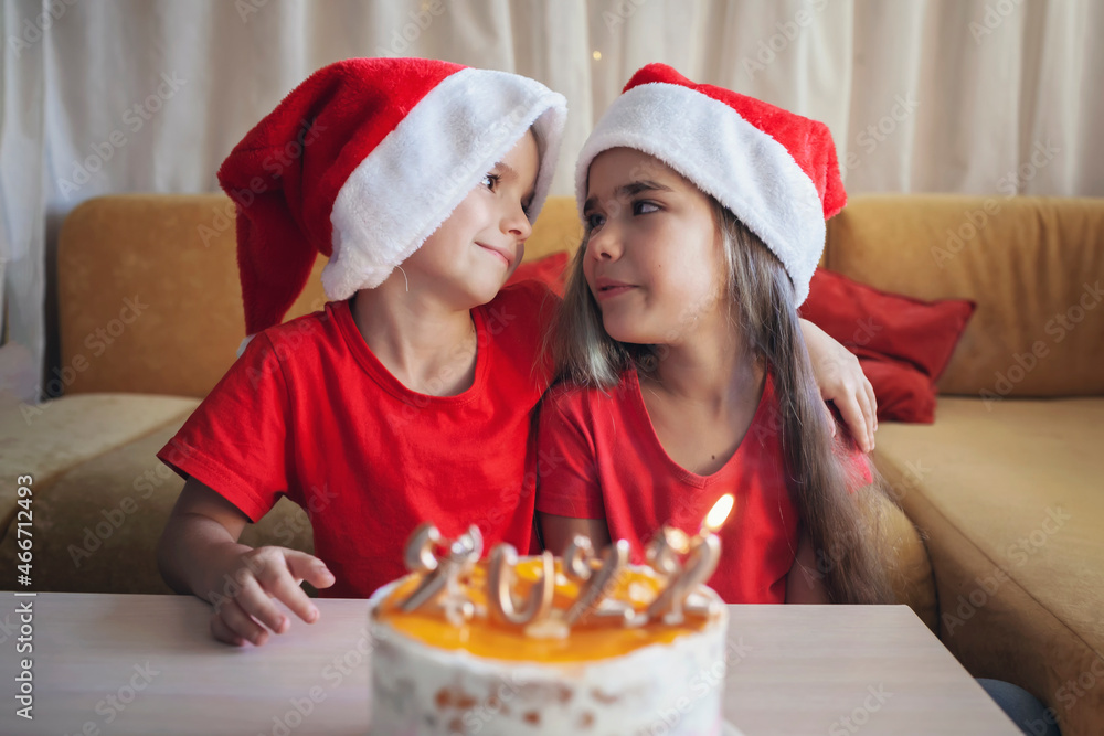 Happy siblings hug each other before Christmas cake with burning golden 2022 candles on top with yellow icing over bokeh lights background, family New Year celebration, focus on kids Happy siblings hug each other before Christmas cake with burning golden 2022 candles on top with yellow icing over bokeh lights background, family New Year celebration, focus on kids