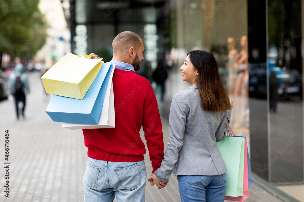 Back view of young international couple walking hand in hand near big mall, holding shopper bags outdoors