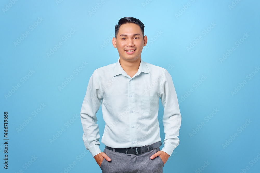 Smiling young handsome businessman smiling at a camera with hands in pockets on blue background Smiling young handsome businessman smiling at a camera with hands in pockets on blue background