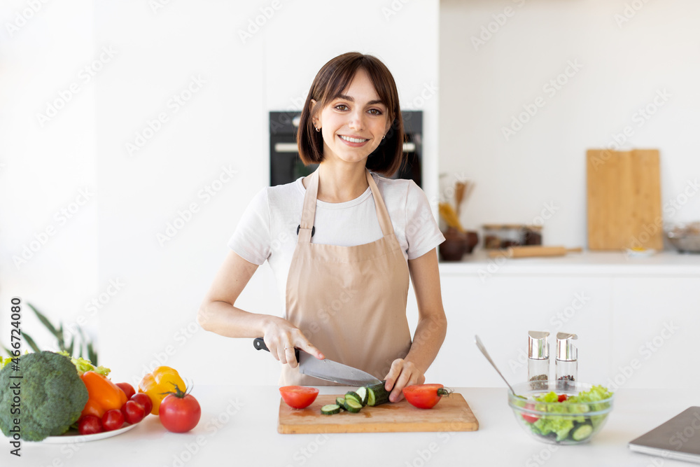 Happy young lady cooking fresh vegetable salad, chopping cucumber and smiling at camera in kitchen
