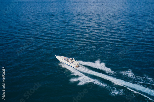 Large open white yacht with people moving fast diagonally on dark blue water aerial view.