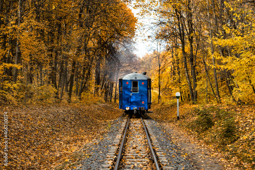 Narrow gauge single track railway and blue vintage departing train in autumn forest in Indian summer, yellow leaves, sunlight and blue sky. Kharkov, Ukraine.