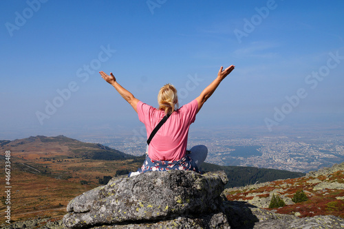 a blonde woman in a pink t-shirt sits on the top of a mountain raising her hands up