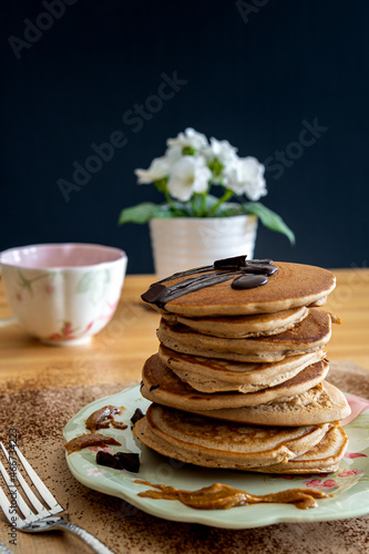 Healthy buckwheat pancake stack with dark chocolate, maple syrup and halvah on a decorative plate. Easy to make gluten free morning breakfast or brunch. Delicious heap of golden pancakes with teacup.